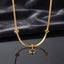 Gold Four-Leaf Clover Monogram Herringbone Chain Pendant Necklace laid flat on white Carrara marble surface in warm natural light showing 18K yellow gold-tone flat herringbone snake chain with round black enamel disc charm quatrefoil star outline charm and hanging round black enamel disc drop pendant with four-petal clover motifs throughout