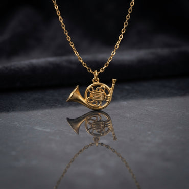 Gold French Horn Music Charm Pendant Necklace flat lay on grey reflective mirror surface with dark navy charcoal satin backdrop showing fine gold cable chain with highly detailed 3D miniature French horn musical instrument charm in matte satin gold tone finish with etched rotary valve detail and mirror reflection below