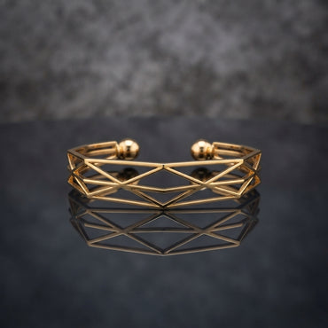Gold Geometric Wireframe Diamond Lattice Open Cuff Bangle in 18K gold-tone PVD stainless steel on dark mirror surface with grey bokeh backdrop showing wide open C-shape cuff with repeating diamond rhombus X-cross geometric lattice wireframe pattern and two smooth gold ball bead finials at open ends with mirror reflection below