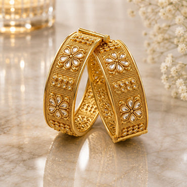 Gold-Tone Wide Granule Kundan Polki Five-Petal Flower Panel Kada Bangle Set of 2 leaning on gold marble surface with crystal candle holder and baby's breath flowers backdrop