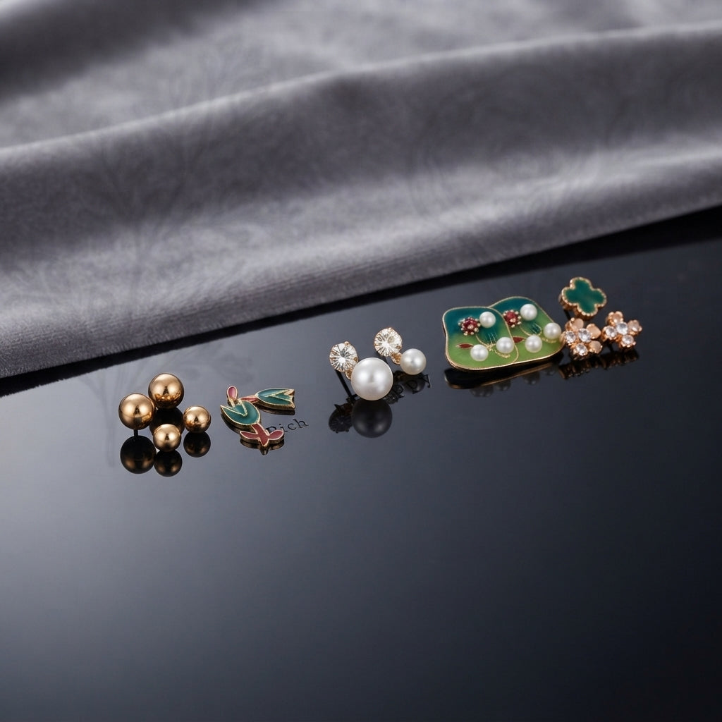 Gold Green Floral Enamel Pearl CZ 4-Pair Stud Earring Set flat lay on black mirror surface with grey fabric backdrop showing gold double ball studs pink coral tulip enamel studs with green leaf white CZ pearl drop studs and large green square enamel stud with pearl row red flower and green clover CZ cluster with mirror reflections below