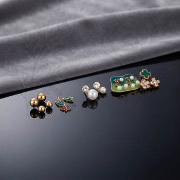 Gold Green Floral Enamel Pearl CZ 4-Pair Stud Earring Set flat lay on black mirror surface with grey fabric backdrop showing gold double ball studs pink coral tulip enamel studs with green leaf white CZ pearl drop studs and large green square enamel stud with pearl row red flower and green clover CZ cluster with mirror reflections below
