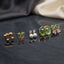 Gold Green Tulip Oval Floral Pearl Clover 5-Pair Stud Earring Set flat lay on dark black stone surface with grey and cream fabric backdrop showing gold ball arc studs green tulip enamel studs with red coral stem white CZ pearl drop studs large green oval enamel stud with pink rose and gold bead border and green clover blush CZ flower cluster stud with mirror reflections below