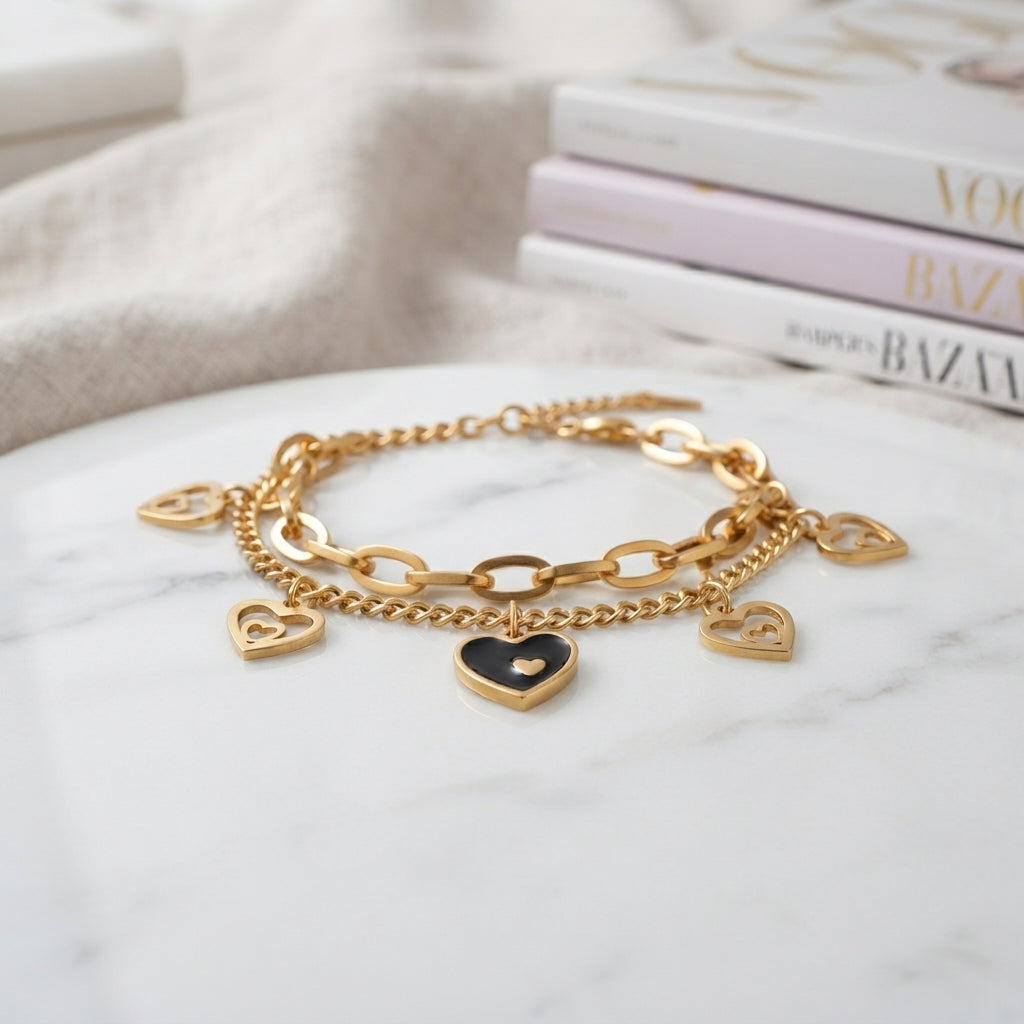 Gold Heart Charm Double Layer Chain Bracelet in 18K gold-tone PVD stainless steel on white marble surface with fashion magazines backdrop showing bold oval paperclip link chain layered with fine curb chain featuring 4 open-cut gold heart outline charms and 1 black enamel filled centre heart charm with small raised gold heart detail