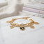 Gold Heart Charm Double Layer Chain Bracelet in 18K gold-tone PVD stainless steel on white marble surface with fashion magazines backdrop showing bold oval paperclip link chain layered with fine curb chain featuring 4 open-cut gold heart outline charms and 1 black enamel filled centre heart charm with small raised gold heart detail