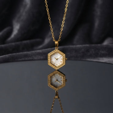 Gold Hexagon Greek Key Watch-Face Pendant Necklace in 18K gold-tone PVD stainless steel on dark grey mirror surface with dark silk backdrop showing bold hexagon pendant with continuous Greek key meander engraved border and decorative white Roman numeral watch-face dial sticker on slim gold cable chain with mirror reflection below