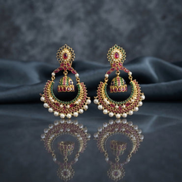 Gold Kundan Meenakari Chandbali Jhumka Earrings in ruby red and emerald green pair on dark mirror surface with grey silk backdrop showing 22K antique gold-tone oval floral Kundan stud with large crescent chandbali frame set with alternating ruby-red and emerald-green teardrop Kundan stones with domed Meenakari jhumka bell and full row of lustrous white pearl drops fringe with mirror reflection below