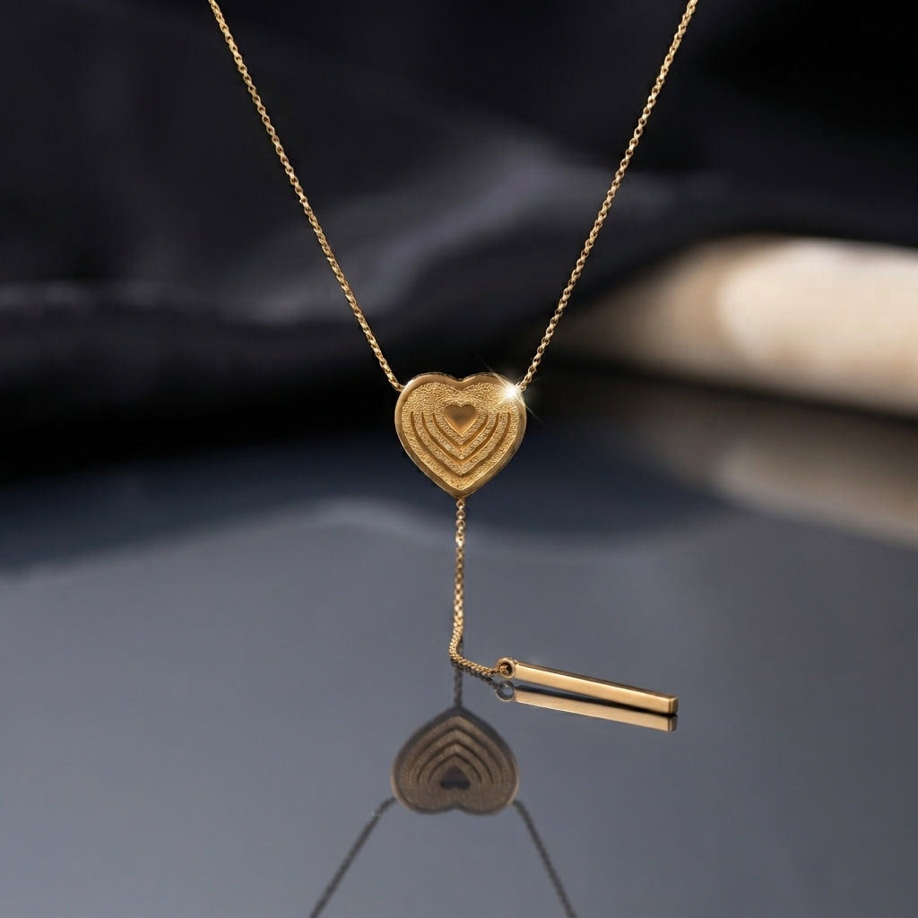 Gold Layered Heart Lariat Bar Drop Necklace flat lay on dark grey mirror surface with dark silk and beige fabric backdrop showing textured concentric layered gold heart pendant on delicate cable chain with Y-drop lariat tail and gold bar charm drop and mirror reflection below