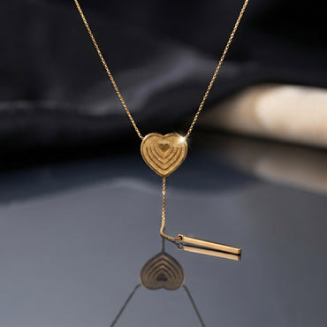 Gold Layered Heart Lariat Bar Drop Necklace flat lay on dark grey mirror surface with dark silk and beige fabric backdrop showing textured concentric layered gold heart pendant on delicate cable chain with Y-drop lariat tail and gold bar charm drop and mirror reflection below