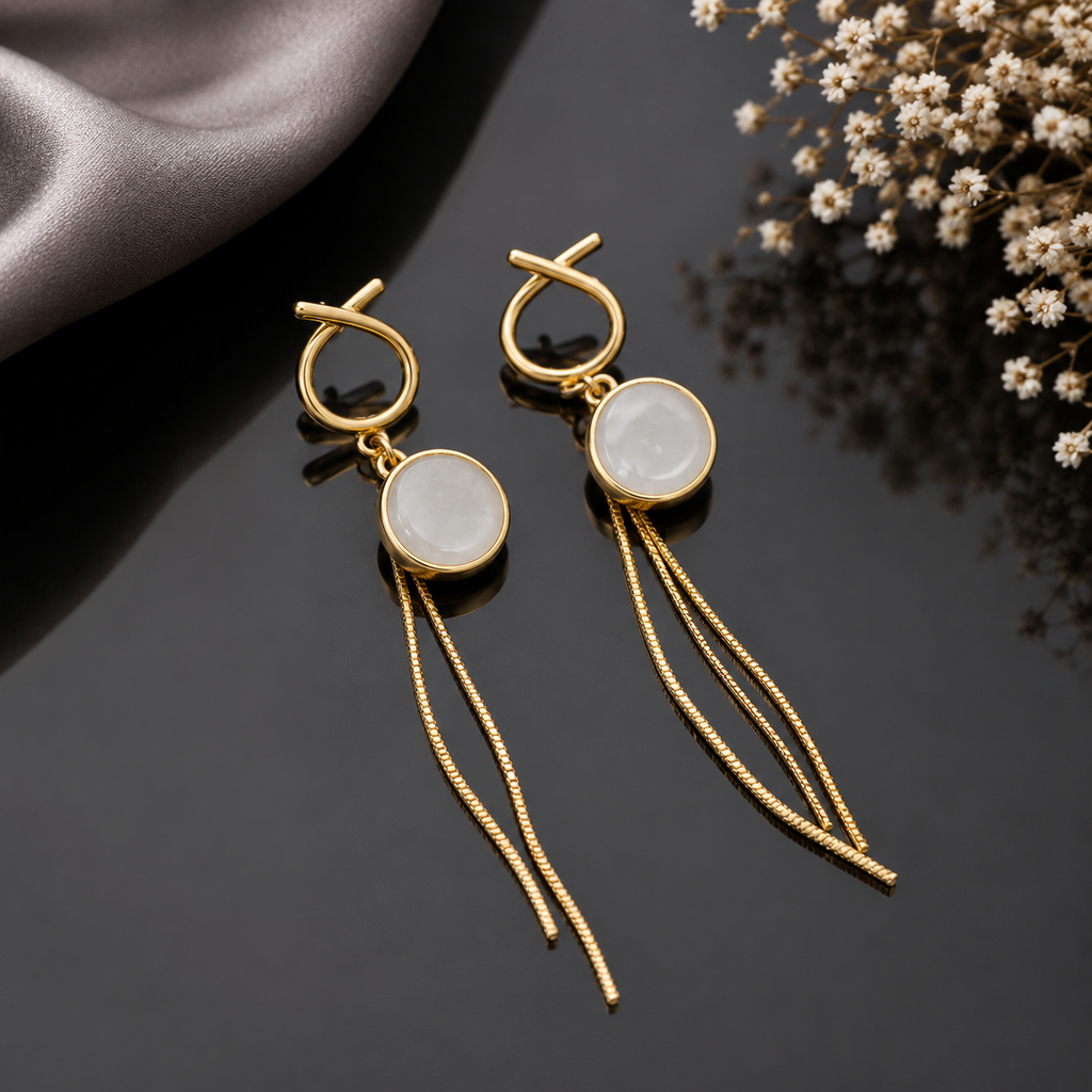 Gold-Tone Loop Knot Top White Moonstone Cabochon Round Bezel & Long Snake Chain Tassel Drop Earrings on black mirror surface with silver satin fabric and dried white baby's breath flowers in background