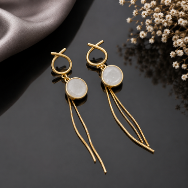 Gold-Tone Loop Knot Top White Moonstone Cabochon Round Bezel & Long Snake Chain Tassel Drop Earrings on black mirror surface with silver satin fabric and dried white baby's breath flowers in background