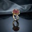 Gold Lotus Flower Cluster CZ Dome Cocktail Ring in Ruby Red — 22K gold-tone lotus flower basket ring with dense ruby-red CZ dome cluster crown, white CZ center accent, white CZ pavé petal halo, and split pavé shank, on dark mirror surface with grey silk backdrop