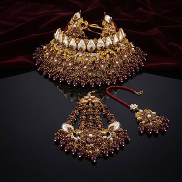 Gold Maroon Ruby Bead and Baroque Pearl Kundan Grand Choker Bridal Set flat lay on dark mirror surface with burgundy velvet backdrop showing 3-piece bridal set with grand wide choker featuring dramatic baroque pearl crown top row and dense antique gold filigree lotus motifs with maroon ruby bead fill and fringe drops with matching large chandbali chandelier earrings and maang tikka with mirror reflection
