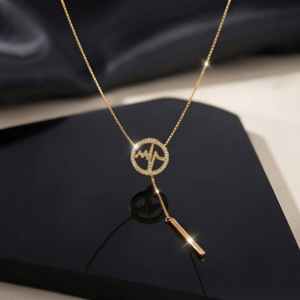 Gold Micro Pavé CZ Heartbeat Circle Lariat Bar Necklace flat lay on black glossy surface with dark silk backdrop showing fully CZ-encrusted open circle pendant with EKG heartbeat wave motif on gold-tone box chain with Y-drop lariat tail and polished gold bar charm drop and mirror reflection below