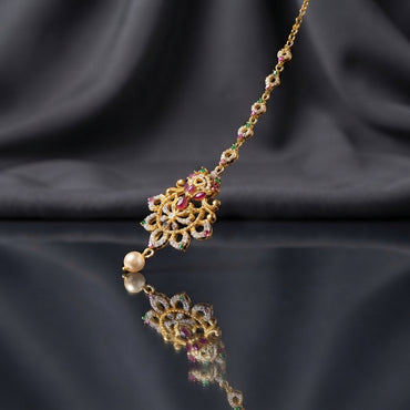 Gold Multi-Color CZ Mughal Floral Fan Maang Tikka with Pearl Drop — antique 22K gold-tone South Asian bridal maang tikka with large fan-shaped Mughal floral pendant featuring ruby CZ center focal marquise ruby CZ accents emerald green CZ details white CZ micro-pavé open fan arches and single cream pearl drop on decorative ring-link CZ chain displayed on dark charcoal satin backdrop with mirror surface reflection