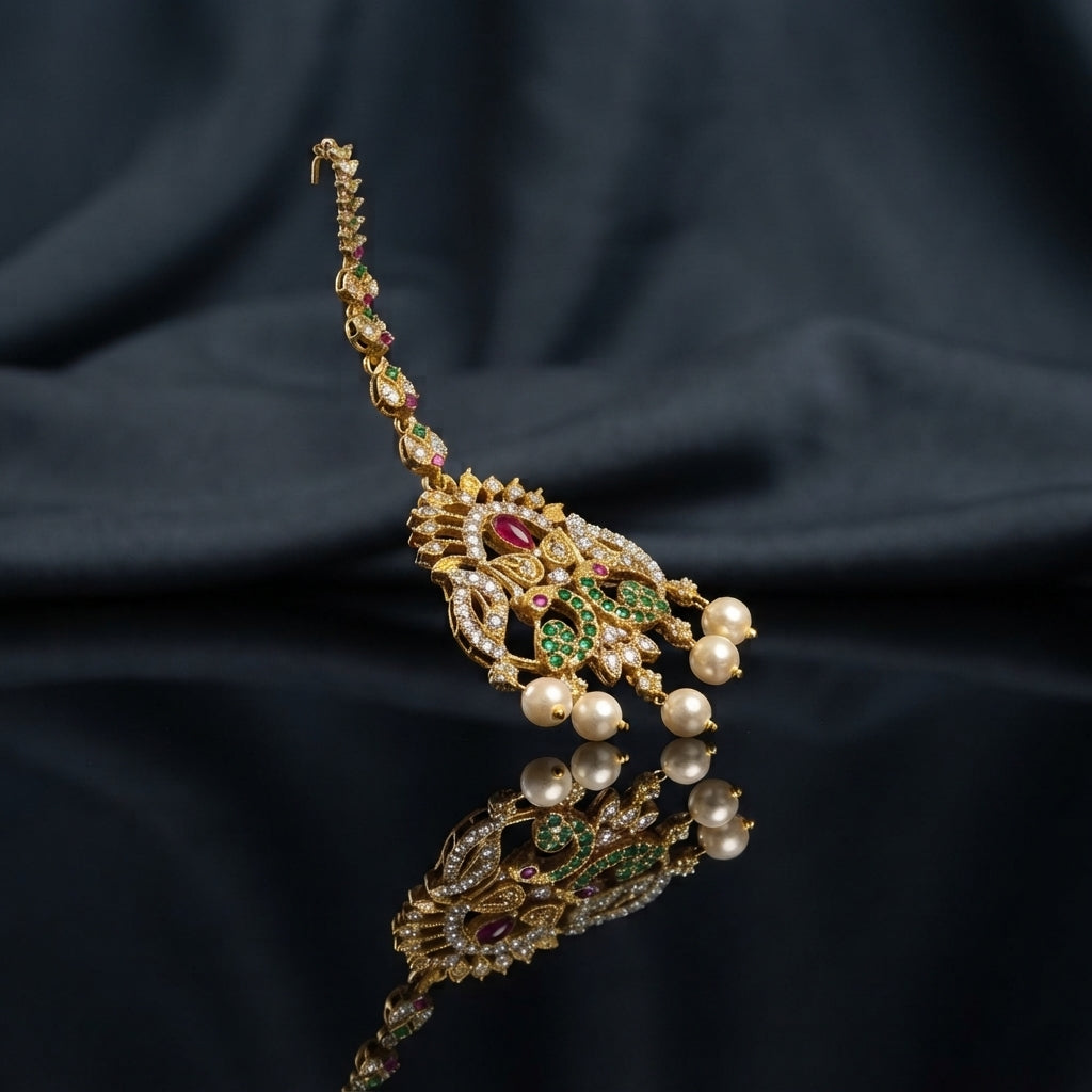 Gold Multi-Color CZ Mughal Peacock Maang Tikka with Pearl Drops — antique 22K gold-tone traditional South Asian bridal maang tikka with elaborate Mughal peacock pendant featuring ruby CZ center focal emerald green CZ peacock wing fill white CZ micro-pavé scrollwork body marquise CZ leaf crown and 3 cream pearl drops on long marquise CZ tikka chain displayed on dark charcoal satin backdrop with mirror surface reflection