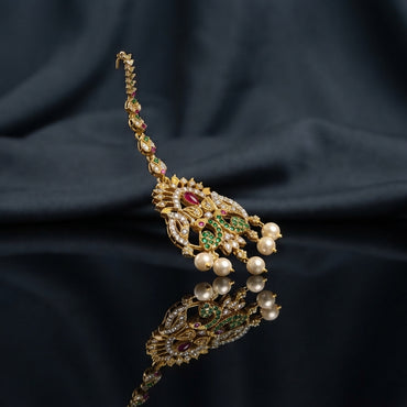 Gold Multi-Color CZ Mughal Peacock Maang Tikka with Pearl Drops — antique 22K gold-tone traditional South Asian bridal maang tikka with elaborate Mughal peacock pendant featuring ruby CZ center focal emerald green CZ peacock wing fill white CZ micro-pavé scrollwork body marquise CZ leaf crown and 3 cream pearl drops on long marquise CZ tikka chain displayed on dark charcoal satin backdrop with mirror surface reflection