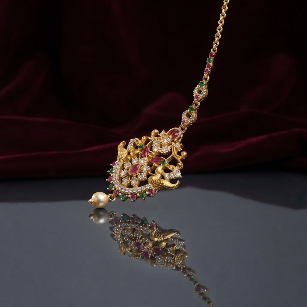 Gold Multi-Color CZ Mughal Twin Peacock Maang Tikka with Pearl Drop — antique 22K gold-tone South Asian bridal maang tikka with large diamond-shaped Mughal twin peacock pendant featuring marquise ruby CZ wing fills emerald green CZ accents white CZ micro-pavé scrollwork and single cream pearl drop on fine gold CZ chain displayed on dark grey mirror surface with burgundy velvet backdrop