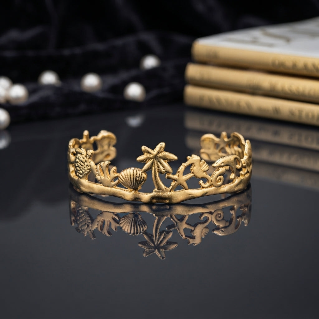 Gold Ocean Marine Life Open Cuff Bangle in 18K antique gold-tone PVD stainless steel on dark grey mirror surface with black silk backdrop and pearl string showing wide open cuff with fully sculpted 3D marine motifs including sea turtle scallop shell palm tree starfish seahorse dolphin coral and wave scrolls with mirror reflection below