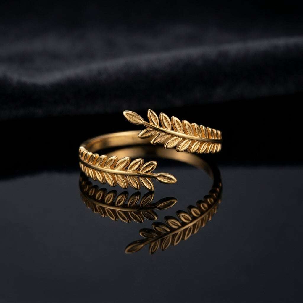 Gold Olive Laurel Leaf Wrap Bypass Adjustable Ring flat lay on dark black mirror surface with dark charcoal navy satin backdrop showing 18K gold-tone open wrap bypass ring with two detailed olive laurel leaf branches with individually raised engraved oval leaves crossing in graceful wrap design with mirror reflection below