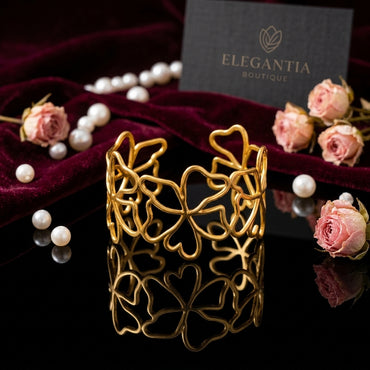 Gold-Tone Openwork Wild Clover Flower & Heart Vine Wide Open Cuff Statement Bracelet on black mirror surface with scattered pearls, pink roses and burgundy velvet backdrop