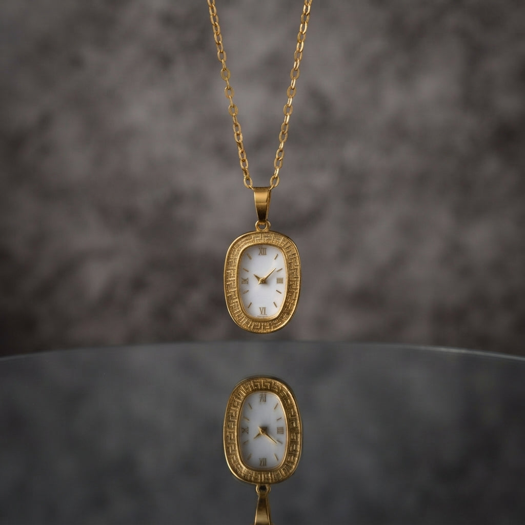 Gold Oval Greek Key Watch-Face Pendant Necklace in 18K gold-tone PVD stainless steel on grey marble surface with taupe bokeh backdrop showing rounded rectangle oval cushion pendant with continuous Greek key meander engraved border and decorative white Roman numeral watch-face dial sticker on slim gold cable paperclip link chain with mirror reflection below