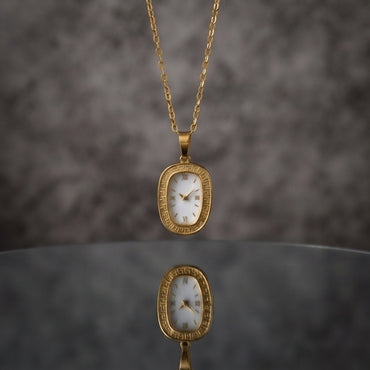 Gold Oval Greek Key Watch-Face Pendant Necklace in 18K gold-tone PVD stainless steel on grey marble surface with taupe bokeh backdrop showing rounded rectangle oval cushion pendant with continuous Greek key meander engraved border and decorative white Roman numeral watch-face dial sticker on slim gold cable paperclip link chain with mirror reflection below