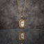 Gold Oval Greek Key Watch-Face Pendant Necklace in 18K gold-tone PVD stainless steel on grey marble surface with taupe bokeh backdrop showing rounded rectangle oval cushion pendant with continuous Greek key meander engraved border and decorative white Roman numeral watch-face dial sticker on slim gold cable paperclip link chain with mirror reflection below