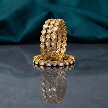 Gold Oval Kundan Polki Full-Bezel Slim Bangle Set of 4 standing upright on dark grey mirror surface with teal silk backdrop showing four matching slim gold bangles fully encircled with continuous oval flat-cut CZ kundan polki stones in antique gold individual bezel settings with mirror reflection below