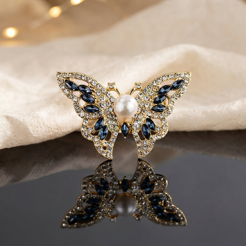 Gold Pearl Butterfly Crystal Brooch in Navy Blue on dark grey mirror surface with cream fabric and warm bokeh backdrop showing large butterfly brooch with open filigree gold wings dense micro-pave white CZ border navy blue marquise crystal wing stones and large faux pearl centre with mirror reflection below