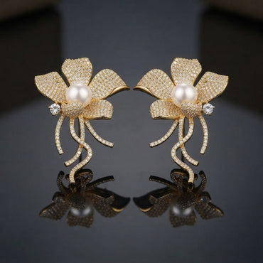 Gold Pearl Pave Orchid Flower Vine Statement Earrings pair on dark grey mirror surface showing large 5-petal orchid magnolia flower fully encrusted in dense micro-pave champagne yellow CZ stones with large round faux pearl cabochon centre white CZ side accent and curling vine tendril stems with full micro-pave CZ draping below in 18K gold-tone plating with mirror reflections below