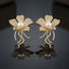 Gold Pearl Pave Orchid Flower Vine Statement Earrings pair on dark grey mirror surface showing large 5-petal orchid magnolia flower fully encrusted in dense micro-pave champagne yellow CZ stones with large round faux pearl cabochon centre white CZ side accent and curling vine tendril stems with full micro-pave CZ draping below in 18K gold-tone plating with mirror reflections below