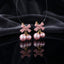 Gold Pink Bow Double Pearl Drop Earrings pair — matte blush pink resin bow studs with double blush rose pink simulated pearl cherry drops on gold links, luxury flat-lay on dark charcoal silk backdrop with mirror reflection