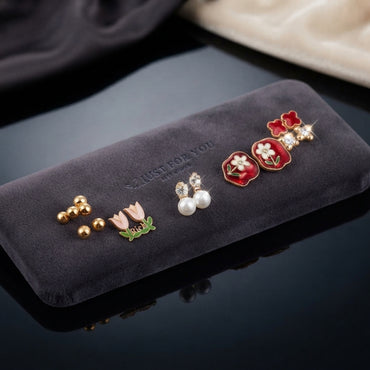 Gold Pink Tulip Red Floral Enamel Pearl CZ 5-Pair Stud Earring Set displayed in open clear Just For You acrylic gift box on black mirror surface with grey and cream fabric backdrop showing gold ball studs pink tulip enamel studs CZ pearl drop studs large red square enamel daisy studs and red clover CZ crystal studs on white velvet insert with mirror reflection below