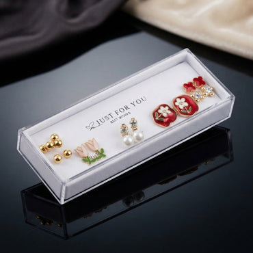 Gold Pink Tulip Red Floral Enamel Pearl CZ 5-Pair Stud Earring Set flat lay on dark grey velvet Just For You tray on black mirror surface with cream fabric backdrop showing gold ball double studs pink blush tulip enamel studs with green leaf white CZ pearl drop studs large red square enamel daisy stud with green leaf and gold border and red clover CZ crystal stud with mirror reflections below
