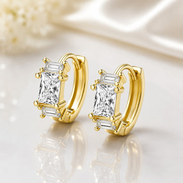 Gold-Tone Princess Cut & Baguette CZ Huggie Hoop Earrings pair on white reflective surface with ivory silk fabric and dried flower backdrop