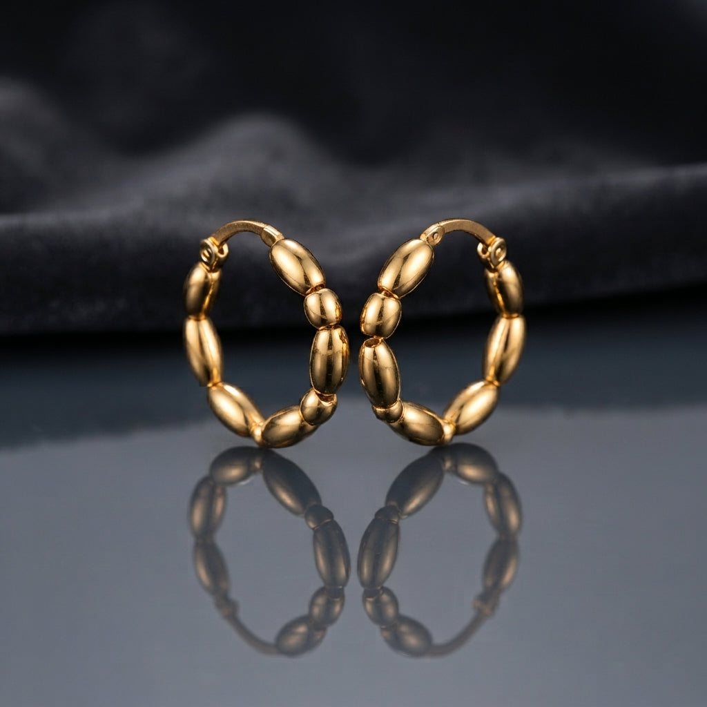 Gold PVD Stainless Steel Chunky Oval Bubble Bead Hoop Earrings pair leaning against each other on dark mirror surface with charcoal slate satin backdrop showing 18K gold PVD coated stainless steel hoops with 7 to 8 large puffy oval barrel beads linked in continuous circle with hinged click closure and sharp mirror reflection below