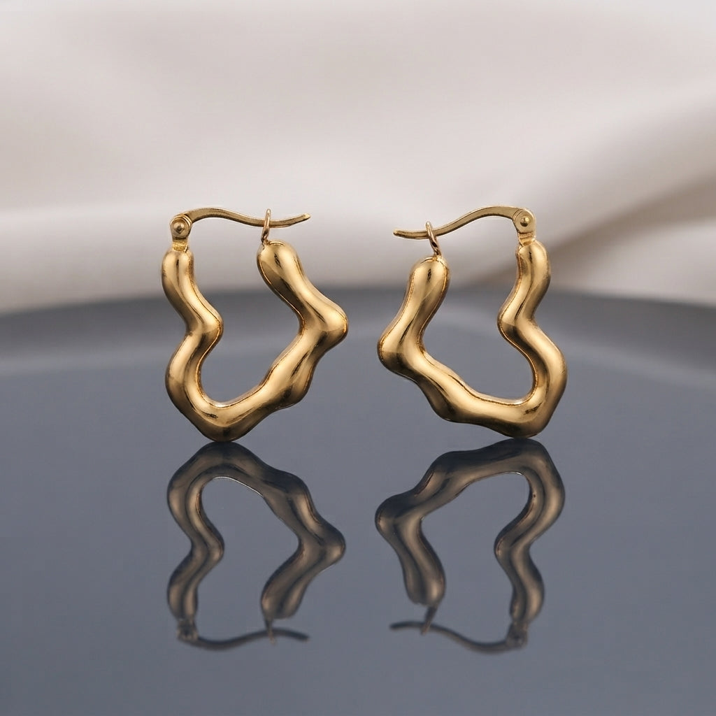 Gold PVD Stainless Steel Organic Wavy Abstract Freeform Hoop Earrings pair standing side by side on dark grey mirror surface with soft warm cream ivory fabric backdrop showing 18K gold PVD coated stainless steel hoops with chunky puffy tube body in organic undulating wavy abstract freeform silhouette satin matte finish with French lever closure and sharp mirror reflection below