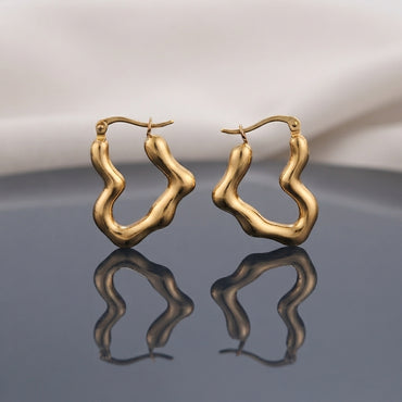 Gold PVD Stainless Steel Organic Wavy Abstract Freeform Hoop Earrings pair standing side by side on dark grey mirror surface with soft warm cream ivory fabric backdrop showing 18K gold PVD coated stainless steel hoops with chunky puffy tube body in organic undulating wavy abstract freeform silhouette satin matte finish with French lever closure and sharp mirror reflection below