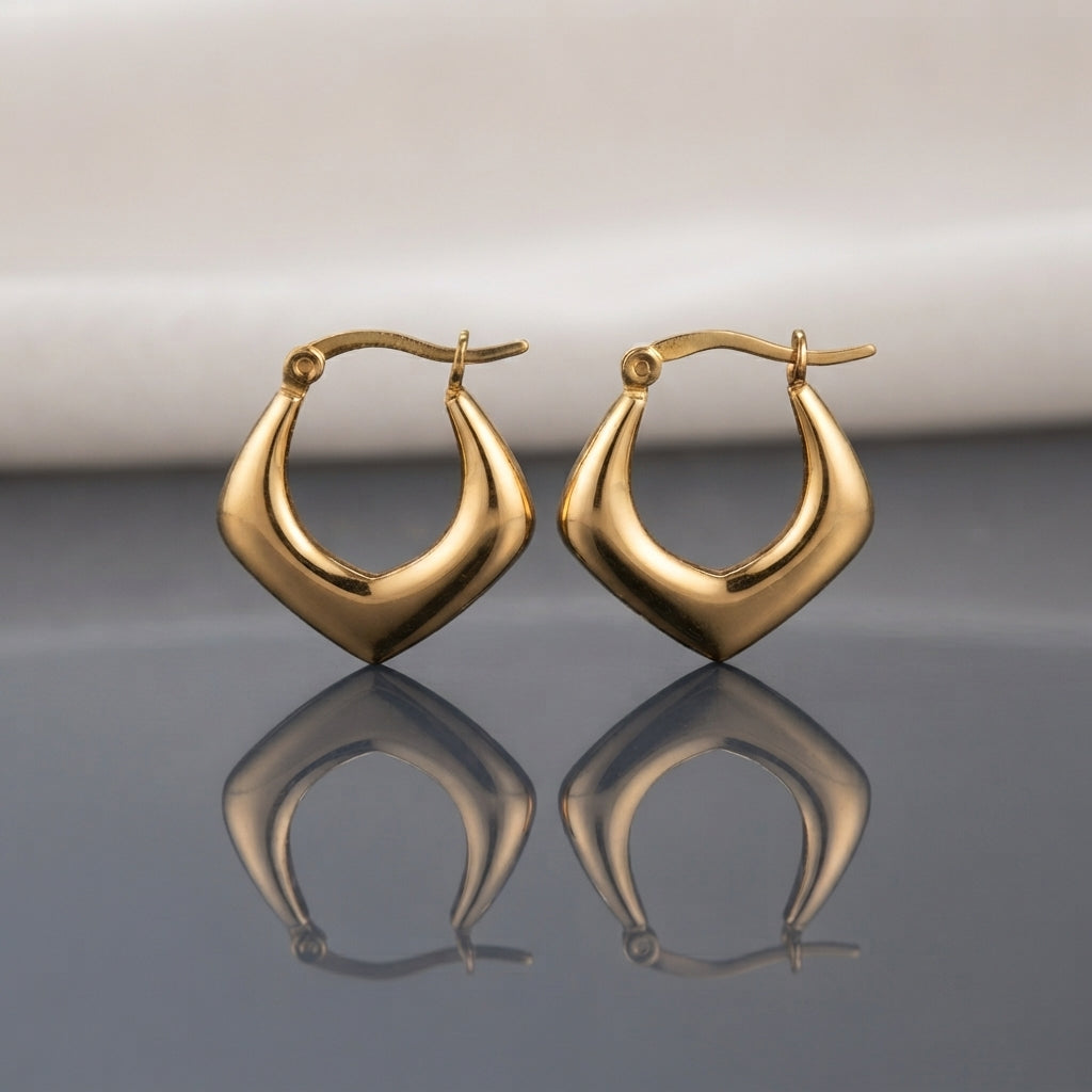 Gold PVD Stainless Steel Puffy Angular Shield V-Point Geometric Hoop Earrings pair standing side by side on light grey mirror surface with soft warm cream ivory fabric backdrop showing 18K gold PVD coated stainless steel hoops with chunky puffy inflated body in wide-bottom shield arrowhead V-point silhouette mirror-polish finish with French lever closure and sharp mirror reflection below