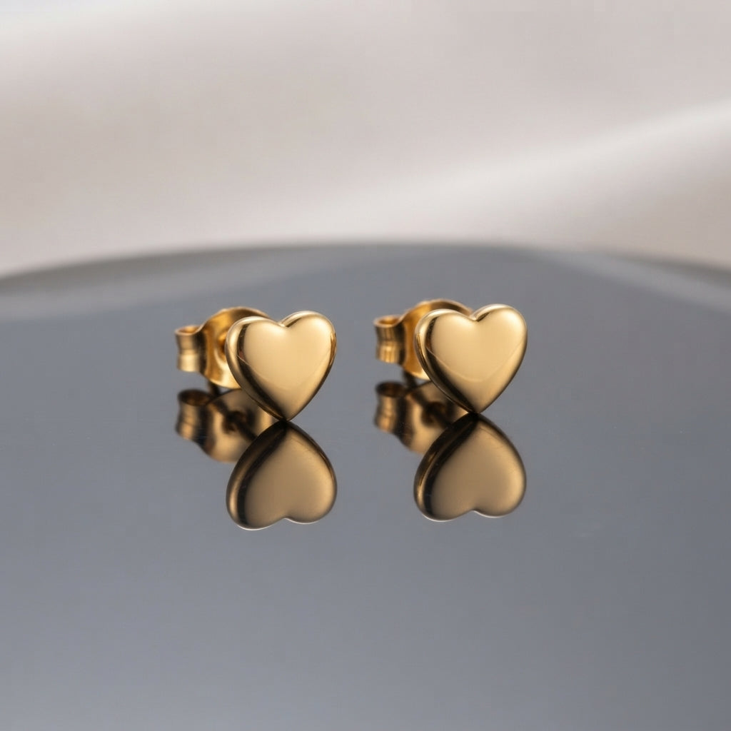 Gold PVD Stainless Steel Smooth Puffy Heart Stud Earrings pair side by side on dark grey ceramic mirror surface with soft warm cream ivory fabric backdrop showing 18K gold PVD coated stainless steel dainty studs with smooth 3D puffy inflated mirror-polish heart face and butterfly push-back post with sharp mirror reflection below