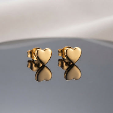 Gold PVD Stainless Steel Smooth Puffy Heart Stud Earrings pair side by side on dark grey ceramic mirror surface with soft warm cream ivory fabric backdrop showing 18K gold PVD coated stainless steel dainty studs with smooth 3D puffy inflated mirror-polish heart face and butterfly push-back post with sharp mirror reflection below