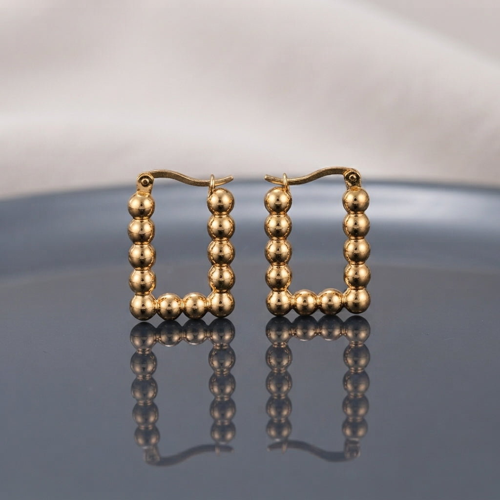 Gold PVD Stainless Steel Square Ball Bead Hoop Earrings pair standing side by side on dark grey ceramic mirror surface with soft warm cream ivory fabric backdrop showing 18K gold PVD coated stainless steel perfect square hoops made of 9 to 10 smooth round mirror-polish gold ball beads with French lever latch-back closure and sharp mirror reflection below