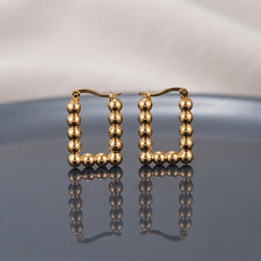 Gold PVD Stainless Steel Square Ball Bead Hoop Earrings pair standing side by side on dark grey ceramic mirror surface with soft warm cream ivory fabric backdrop showing 18K gold PVD coated stainless steel perfect square hoops made of 9 to 10 smooth round mirror-polish gold ball beads with French lever latch-back closure and sharp mirror reflection below