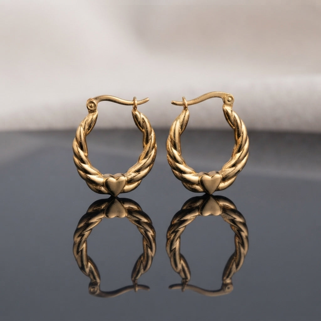 Gold PVD Stainless Steel Twisted Rope Hoop Earrings with Heart Accent pair standing side by side on dark grey mirror surface with soft warm cream ivory fabric backdrop showing 18K gold PVD coated stainless steel hoops with two thick satin-brushed twisted rope braided strands and smooth puffy mirror-polish 3D heart accent at base with French lever closure and sharp mirror reflection below
