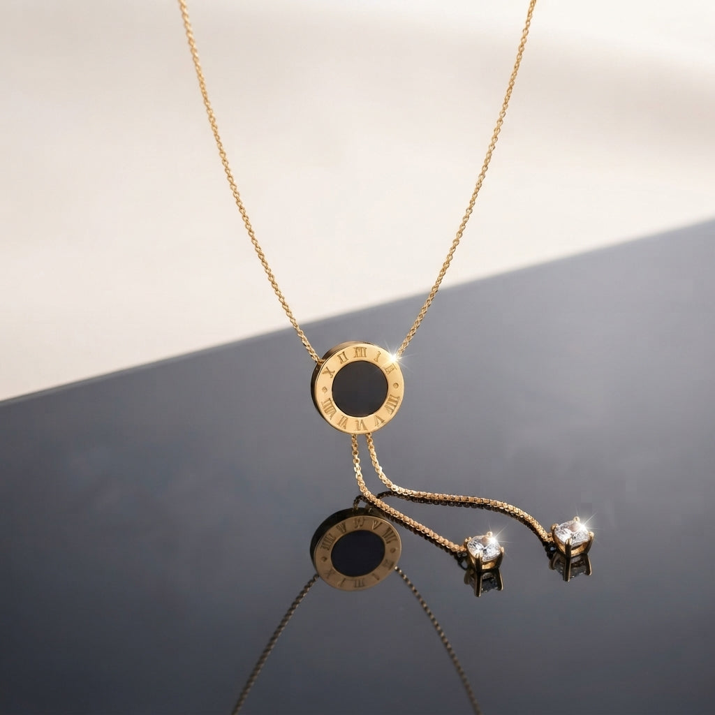 Gold Roman Numeral Circle Enamel Lariat Necklace in black enamel flat lay on grey and white split surface with gold cable chain showing circular Roman numeral engraved pendant with black enamel center and double drop lariat chain with cushion CZ bezel drops and mirror reflection below