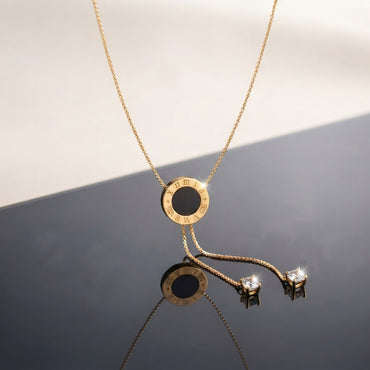 Gold Roman Numeral Circle Enamel Lariat Necklace in black enamel flat lay on grey and white split surface with gold cable chain showing circular Roman numeral engraved pendant with black enamel center and double drop lariat chain with cushion CZ bezel drops and mirror reflection below