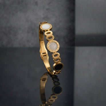 Gold Roman Numeral CZ Pave Triple Disc Bangle in 18K gold-tone plating on dark grey mirror surface showing slim rigid open oval link band bangle with white enamel disc at top CZ pave dome with Roman numeral engraved gold border at centre and black onyx disc at bottom with push-button fold-over clasp and mirror reflection below