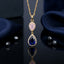 Gold-Tone Rose Pink & Sapphire Blue CZ Double Teardrop Halo Drop Pendant Necklace on black mirror surface with dark navy blue velvet backdrop and scattered black faceted beads