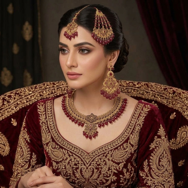 Gold-Tone Ruby Red Bead Filigree Choker & Chandbali Bridal Jewelry Set worn on Pakistani female bridal model in deep maroon velvet lehenga seated on ornate bridal chair with burgundy curtain backdrop